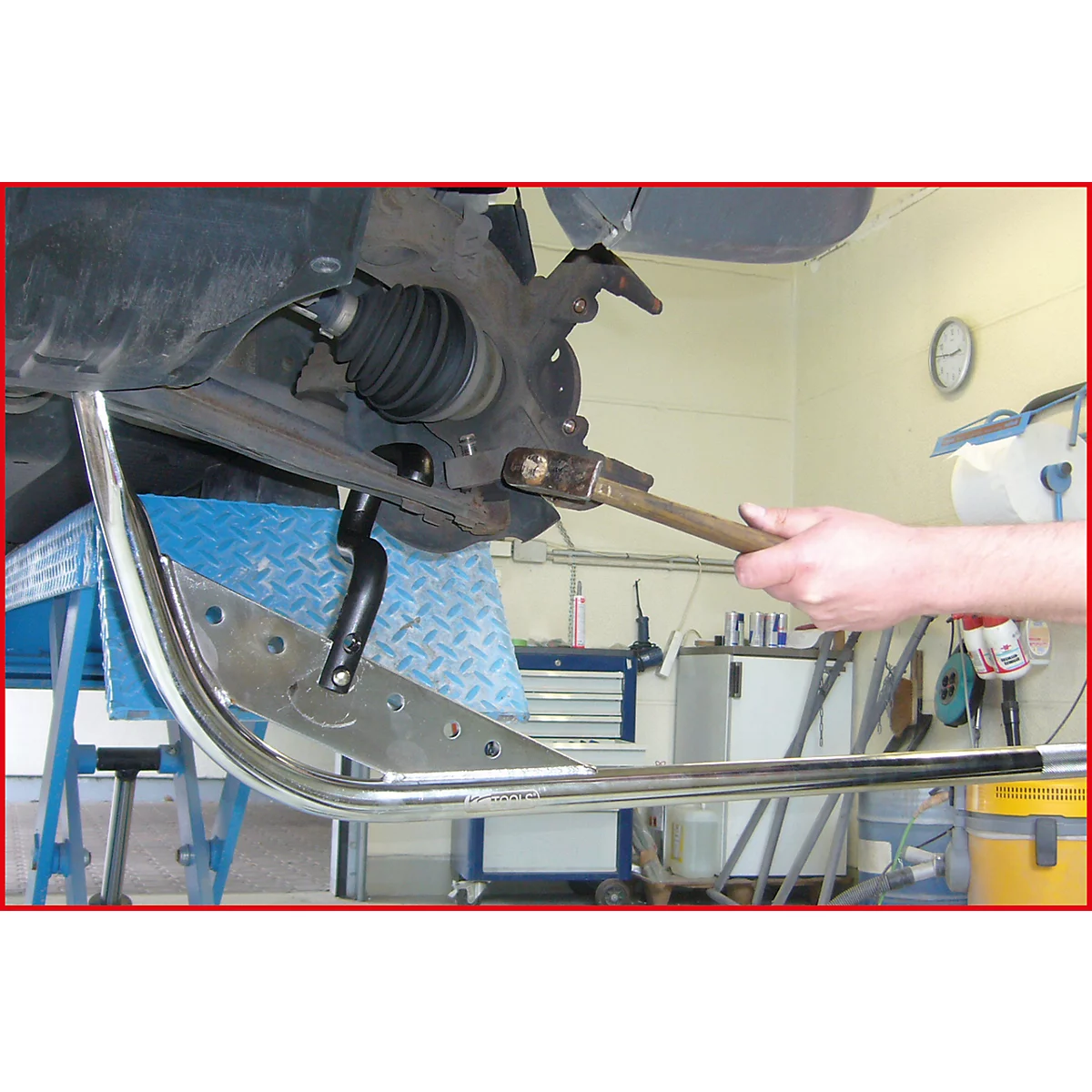 Ein Mechaniker schlägt mit einem Hammer auf ein Autoteil, während eine silberne Stütze das Fahrzeug von unten hält.